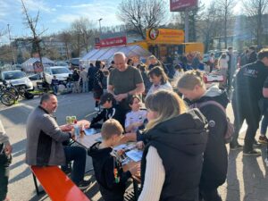 Familien sammeln StickerStars-2026-Sticker an Tischen auf dem Parkplatz vor dem REWE-Markt