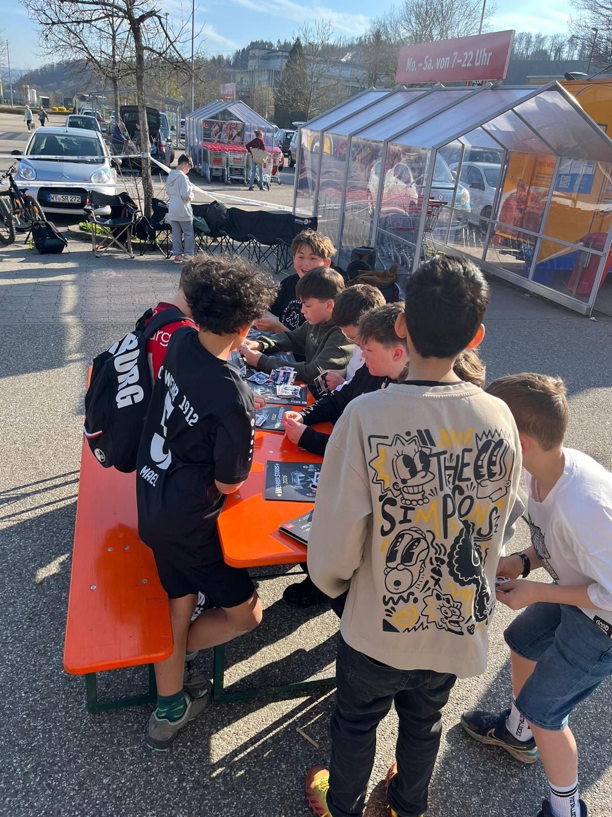 Kinder sitzen zusammen und sammeln StickerStars-2026-Sticker auf dem Parkplatz vor dem REWE-Markt