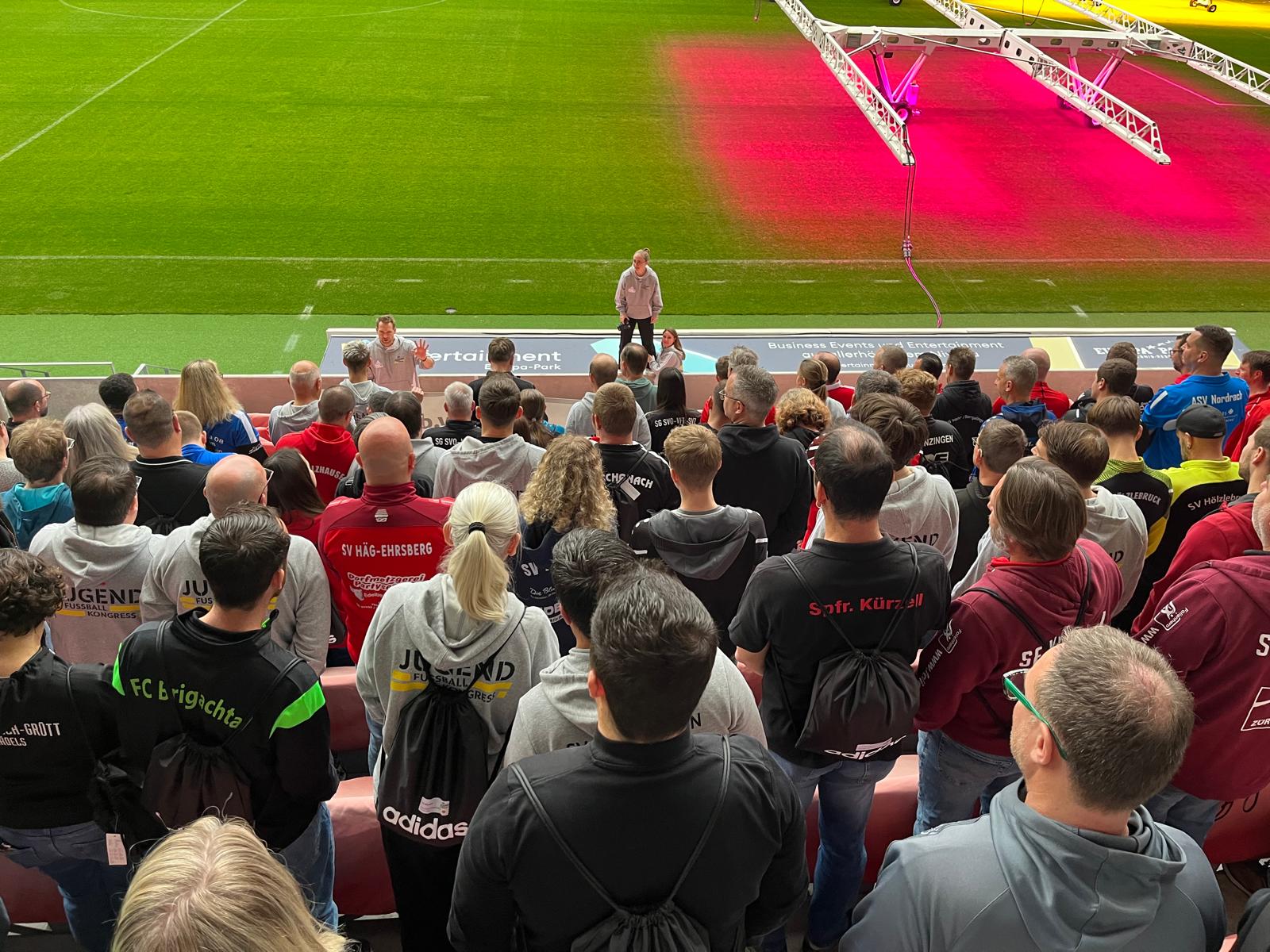 Referent spricht zu einer großen Gruppe von Trainern und Vereinsvertretern auf dem Spielfeld des Europa-Park Stadions