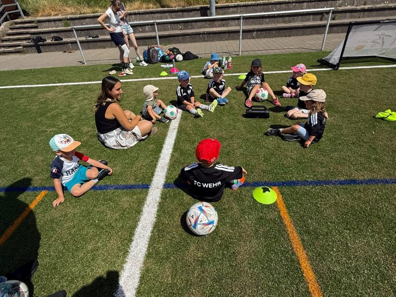 Kinder der Mini Ballschule (U5) sitzen mit ihren Trainerinnen auf dem Rasen beim Training